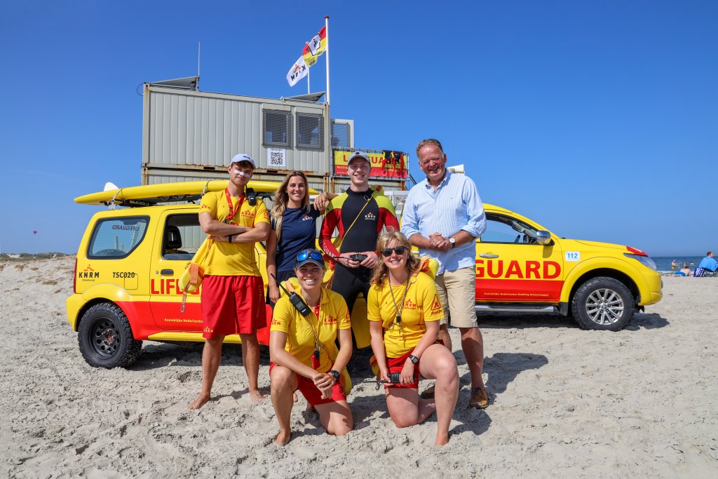 Burgemeester Roel Cazemier samen met vier lifeguards en coördinator Maud Muller op het strand van Terschelling, met op de achtergrond de lifeguard-auto's en post.