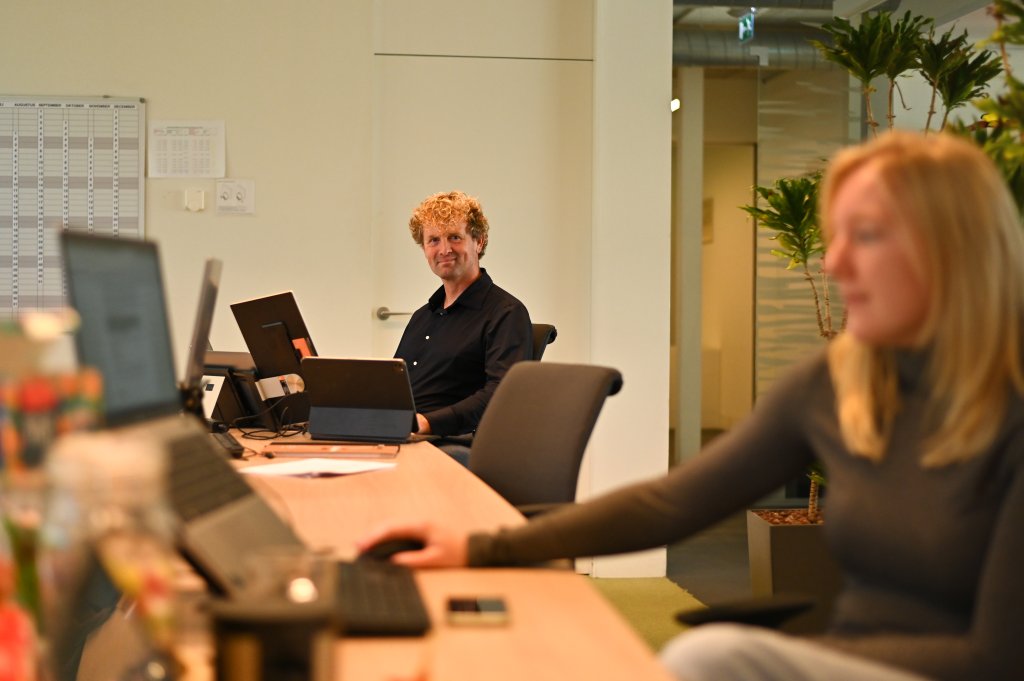Edzart zit aan tafel achter zijn laptop en kijkt de camera in, op de voorgrond rechts is een collega vaag in beeld.