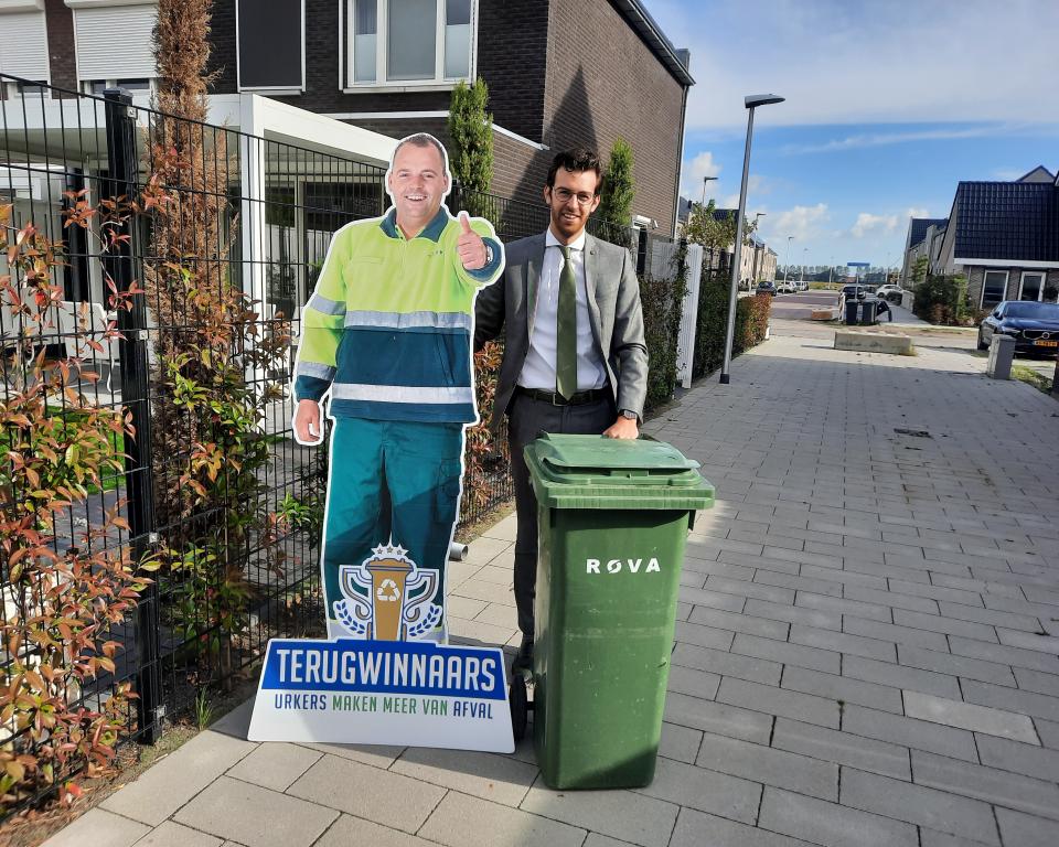 Wethouder Nathanaël Middelkoop met een GFT-afvalbak en een kartonnen afbeelding van een vuilnisman met bord terugwinnaars, Urkers maken meer van afval