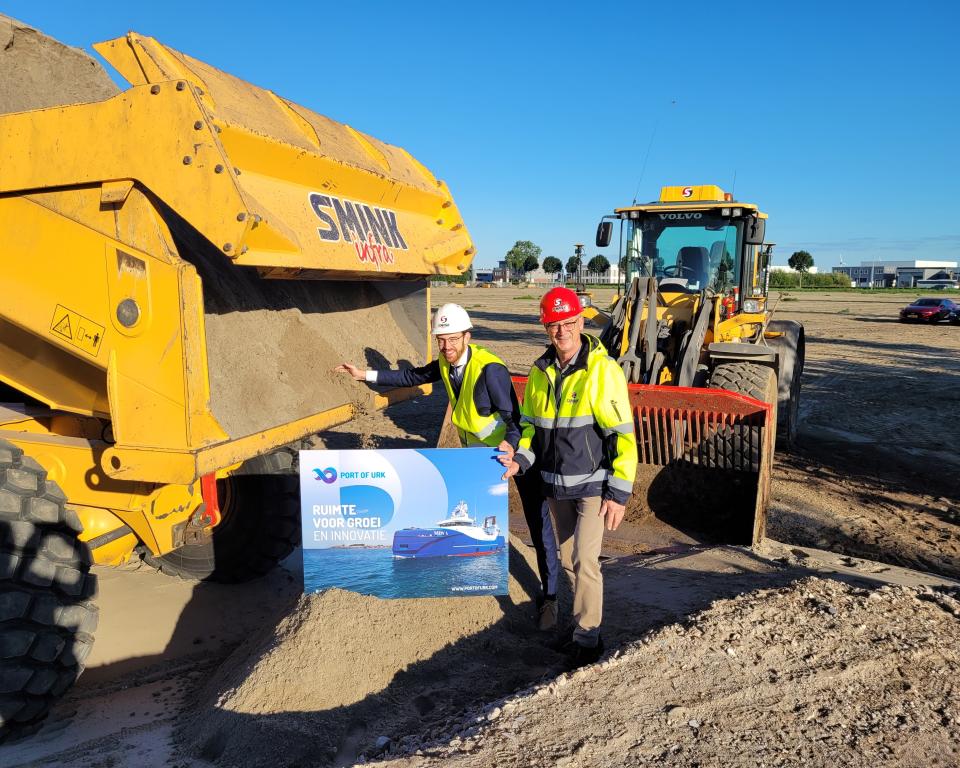 Wethouder Middelkoop en Henny Smink staan voor een shovel met een veiligheidshelm op hun hoofd..