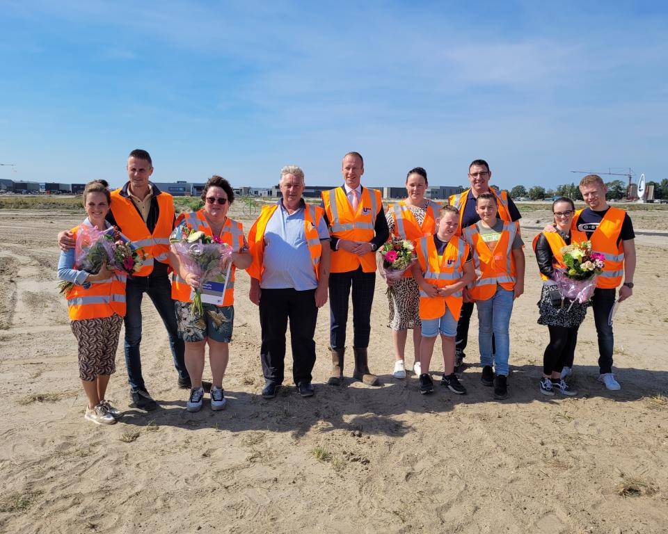 De eerste kopers van een kavel in de Zeeheldenwijk staan in het zand samen met burgemeester Van den Bos. Ze hebben allemaal een oranje hesje aan en de kopers hebben een bos bloemen gekregen.