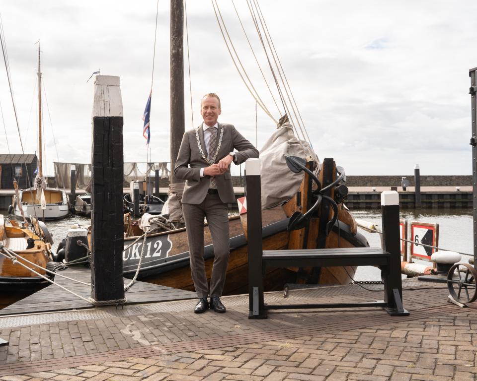 Burgemeester Cees van den Bos staat voor een houten schip met de naam UK 12 in de haven van Urk