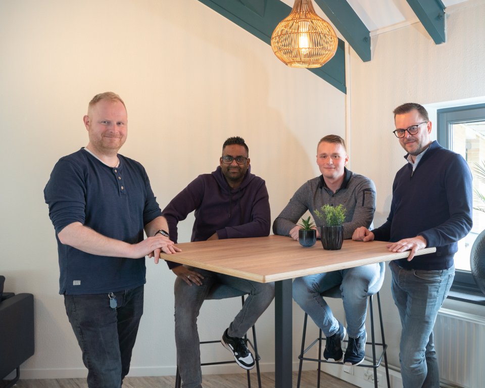 Twee mannen staan en twee mannen zitten achter een tafel