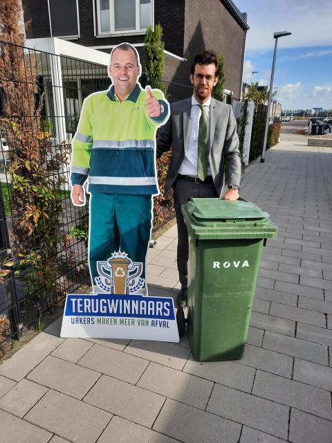 Wethouder Nathanaël Middelkoop bij een GFT-container en een kartonnen bord van een vuilnisman met een bordje terugwinnaars, Urkers maken meer van afval