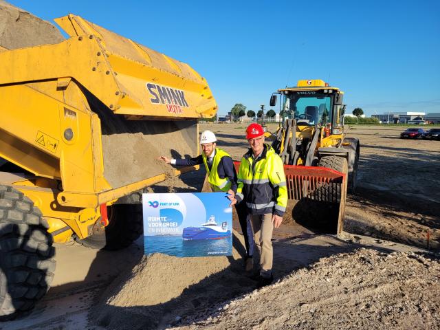 Wethouder Middelkoop en Henny Smink staan voor een shovel met een veiligheidshelm op hun hoofd..