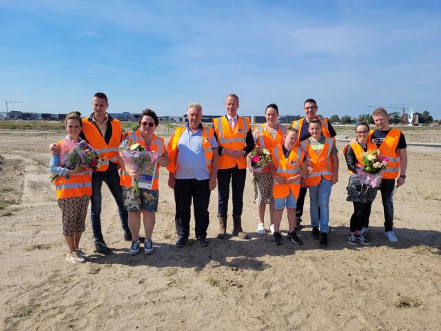 De eerste kopers van een kavel in de Zeeheldenwijk staan in het zand samen met burgemeester Van den Bos. Ze hebben allemaal een oranje hesje aan en de kopers hebben een bos bloemen gekregen.