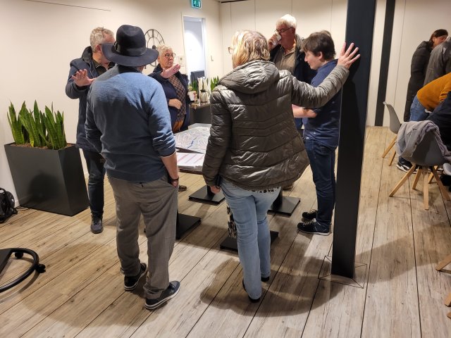 een foto van één inloopbijeenkomst, op de foto staan 6 mensen rond een tafel met elkaar in gesprek. 