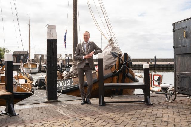 Burgemeester Cees van den Bos staat voor een houten schip met de naam UK 12 in de haven van Urk