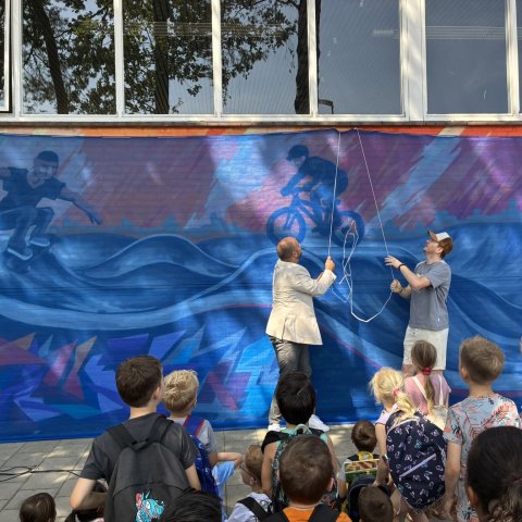 Kinderen kijken toe terwijl twee personen een doek onthullen voor een grote muurschildering op een schoolgebouw met afbeeldingen van een skateboarder en een fietser.