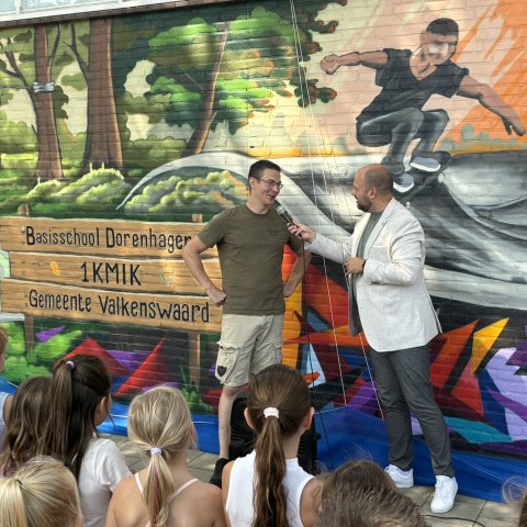 Twee personen staan voor een kleurrijke muurschildering op een schoolplein, terwijl één persoon de ander interviewt met een microfoon. Op de voorgrond kijken kinderen toe. De muurschildering bevat een boslandschap, een skateboarder en de tekst "Basisschool Dorenhagen".