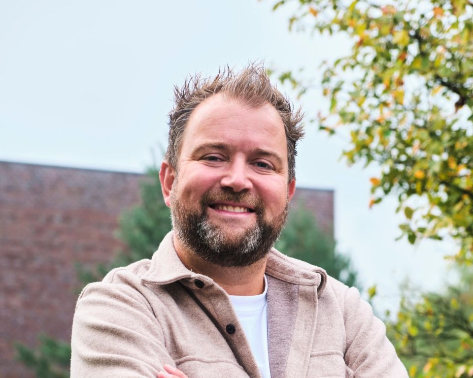 Stadsdeelmanager Baer, staand met armen over elkaar voor een gebouw met groen