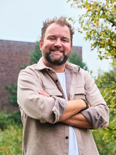 Stadsdeelmanager Baer, staand met armen over elkaar voor een gebouw met groen