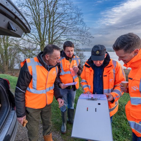 4 mannen met oranje jassen of hesjes buigen zich over een map met informatie. Ze staan buiten bij een geopende achterklep van een auto. 