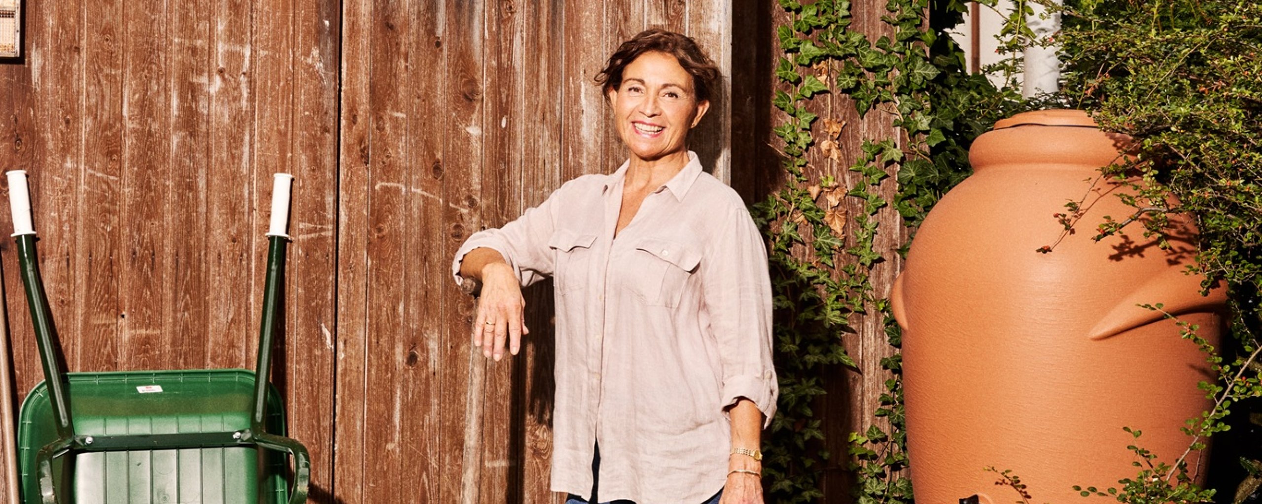 Een vrouw staat met een schep in haar hand bij een schuurtje. Rechts van haar staat een regenton, met daaronder een gieter.