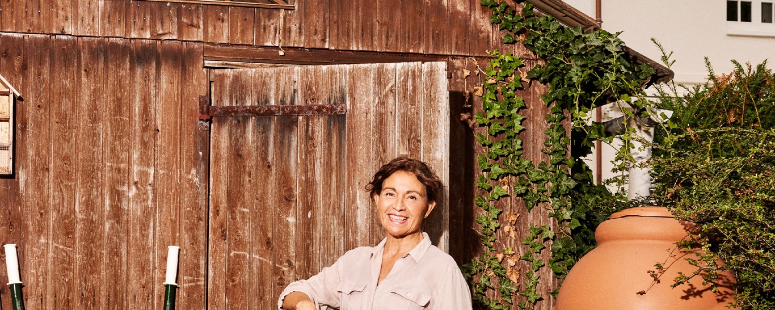 Een vrouw staat met een schep in haar hand bij een schuurtje. Rechts van haar staat een regenton, met daaronder een gieter.