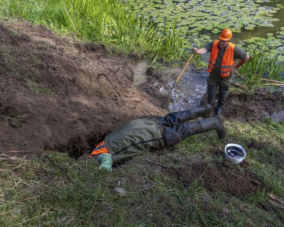 Twee mannen beverhol. Een van de  mannen staat, de andere ligt en verdwijnt met zijn hoofd en deel bovenlichaam in het hol. 