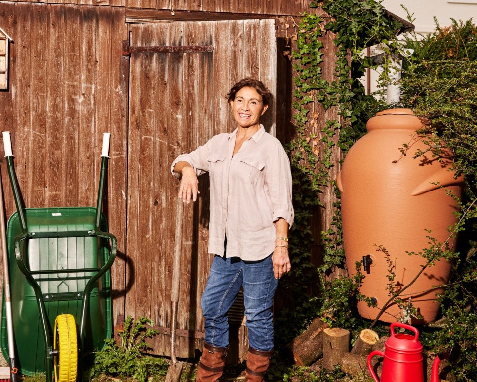 Een vrouw staat met een schep in haar hand bij een schuurtje. Rechts van haar staat een regenton, met daaronder een gieter.