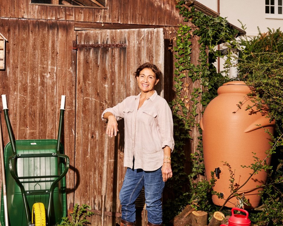 Een vrouw staat met een schep in haar hand bij een schuurtje. Rechts van haar staat een regenton, met daaronder een gieter.