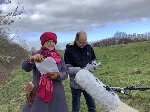 Ineke Knoeff in gesprek met Henk van Dijk terwijl ze buiten in het gras lopen met een papier in de hand en een grote microfoon die hen wordt voorgehouden