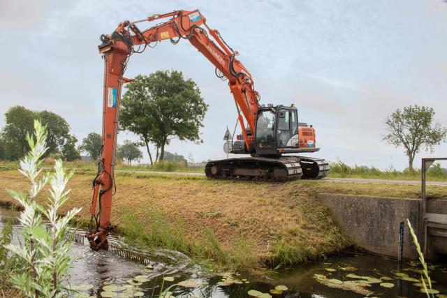 Een kraan langs de waterkant met een grote arm met maaier in de sloot.