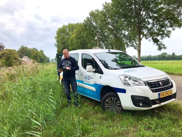 Maarten met een tablet in de hand langs een watergang met veel begroeiing. De waterschapsauto staat achter hem