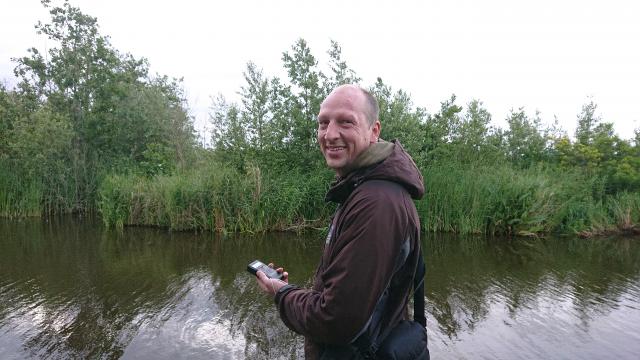 Stefan van Lieshout met detector in de hand, langs een kanaal met planten op de achtergrond.