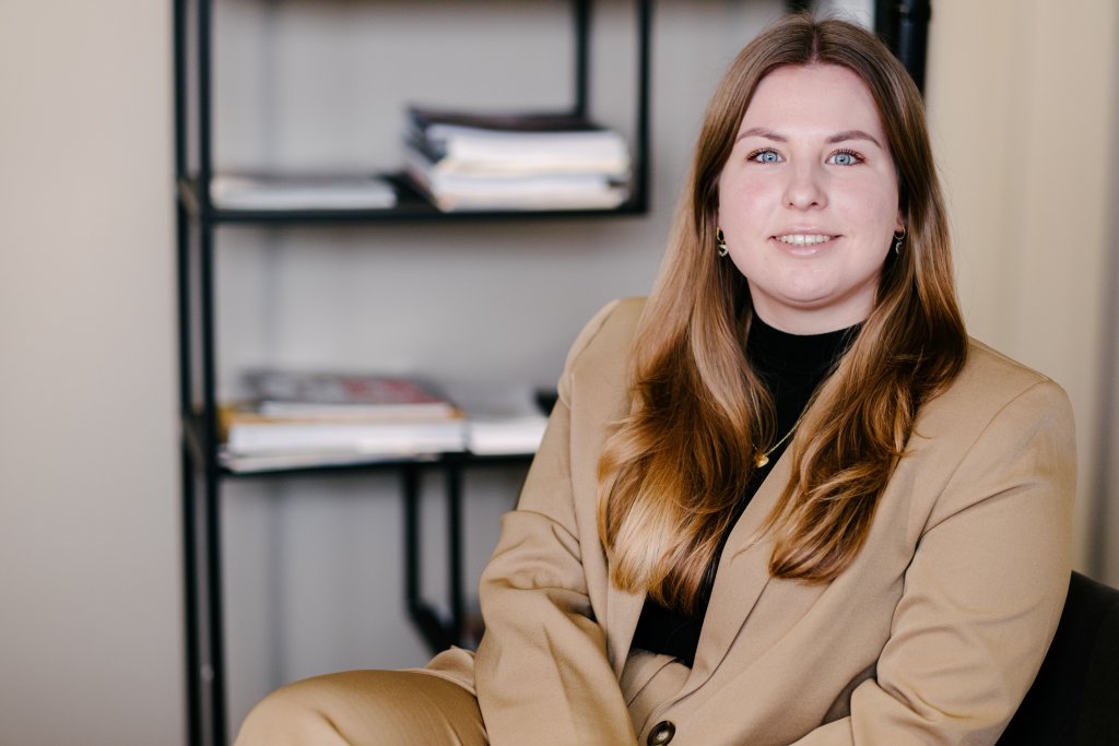 Donkerblonde dame zittend in een kantoorsetting, met kast met papieren op de achtergrond.