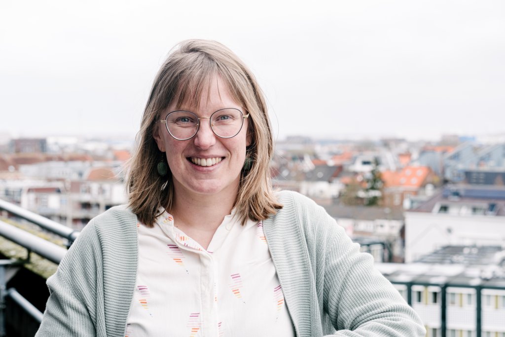 Lachende donkerblonde vrouw met bril, met stad van bovenaf als achtergrond.