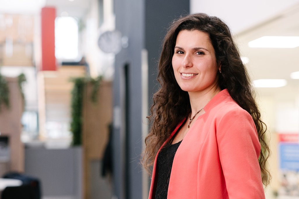 Vrouw met lange krullende donkerbruine haren, kantoorruimte als achtergrond.