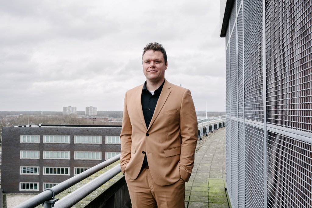 Man met donker haar in lichtbruin pak, op de achtergrond Bergen op Zoom.