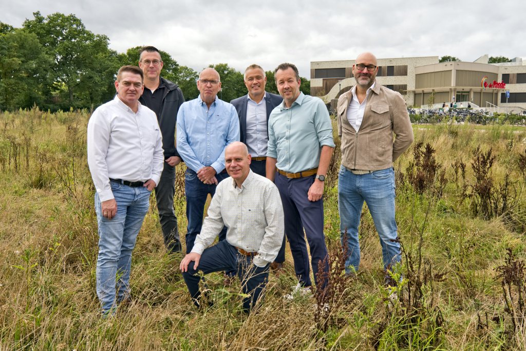 Zeven mannen staan samen in een grasveld bij het toekomstige terrein van sporthal Esrand, met sportcentrum De Passie op de achtergrond.