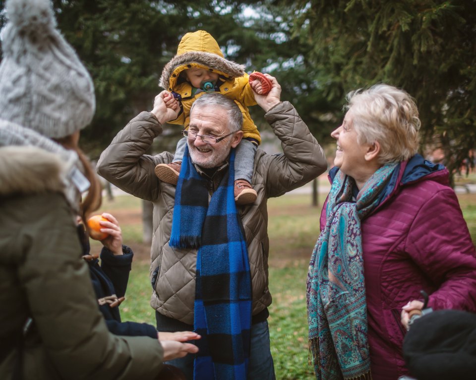 Een peuter zit op de schouders van een oudere man. Naast hem staat een oudere vrouw. Ze lachen.
