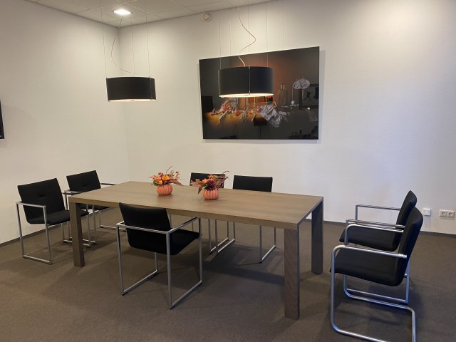 De Victoriakamer in trouwopstelling. Er staat een rechthoekige bruine tafel met daaromheen 7 zwarte stoelen. Boven de tafel hangen 2 zwarte lampen. Op tafel staan 2 oranje bloemstukken. Aan de muur hangt een kunstwerk.