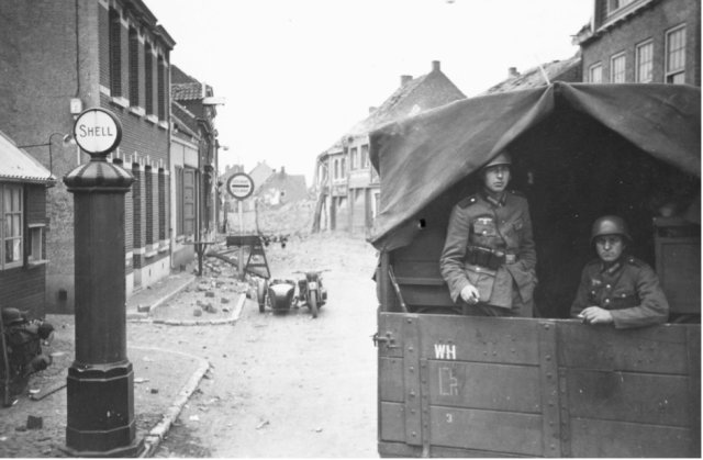 Duitsers van een verbindingseenheid poseren vanuit een vrachtwagen. Op de achtergrond is de ravage te zien van het kruispunt wat kort daarvoor door het Belgische leger werd opgeblazen. Mei 1940. 