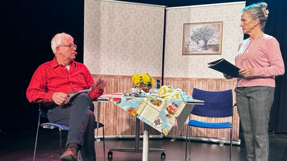 Foto uit de theatervoorstelling De Dementie van Jet en Harrie. Op het toneel zit een man in een stoel een tijdschrift te lezen, een vrouw staat ernaast met een mapje papieren in haar handen.