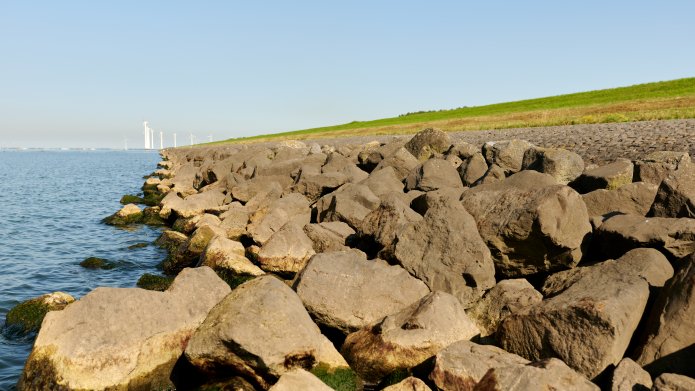 De IJsselmeerdijk met stenen op de voorgrond, water aan de zijkant en een strook gras aan de rechterzijde. 