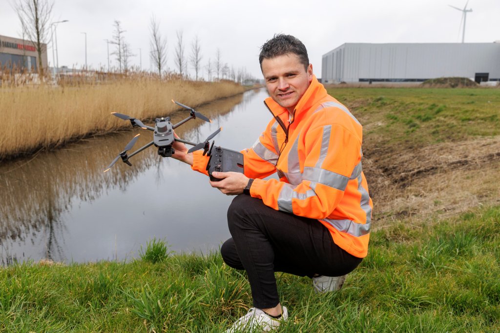 Man in werkjas houdt drone en afstandsbediening vast en zit bij een sloot. 