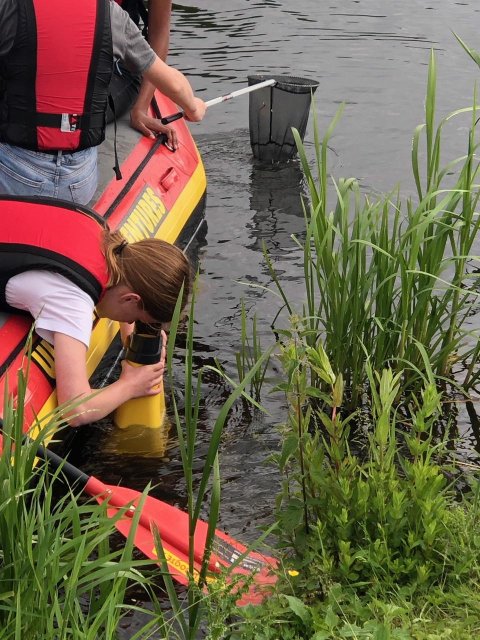 Een scholier zit in een opblaasboot (raft) op het water en kijkt met een geel apparaat in het water. Voor haar zit een klasgenoot met een schepnet. 