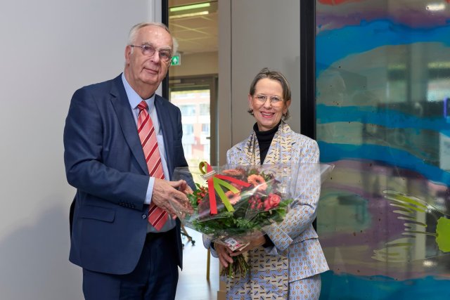 Man en vrouw staan in een ruimte. Vrouw heeft bos bloemen vast. 