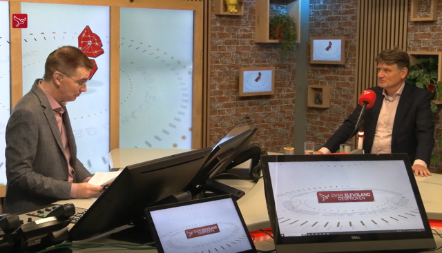 Twee mannen in een televisiestudio aan een tafel met microfoons en beeldschermen