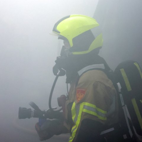 Brandweerpersoon van opzij gezien in volledige uitrusting met helm en gezichtsmasker in de rook tijdens de oefening