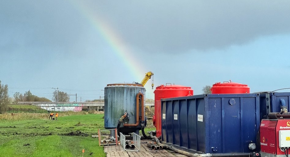 Containers geplaatst rondom de gegraven put bij de bomruiming, op de achtergrond een halve regenboog