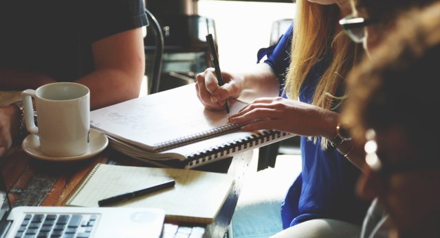 Groep mensen aan een tafel in overleg, kopje koffie op tafel, notitieblok en laptop erbij