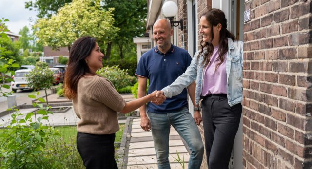 Twee vrouwen geven elkaar de hand voor de deur van een huis, 1 van de vrouwen is de bewoonster en staat in de deuropening met naast haar een man
