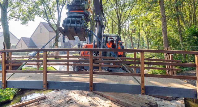 Wethouder Wybe Zijlstra helpt mee met het plaatsen van een nieuwe brug met hulp van een graafmachine