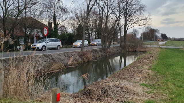 Drie auto's achter elkaar rijdend op een boerenlandweg, op de voorgrond een sloot en op de achtergrond een witte boerderij met rietkap