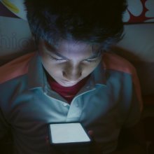 Een tiener zit in een donkere kamer op zijn telefoon. Het felle scherm schijnt in zijn gezicht.